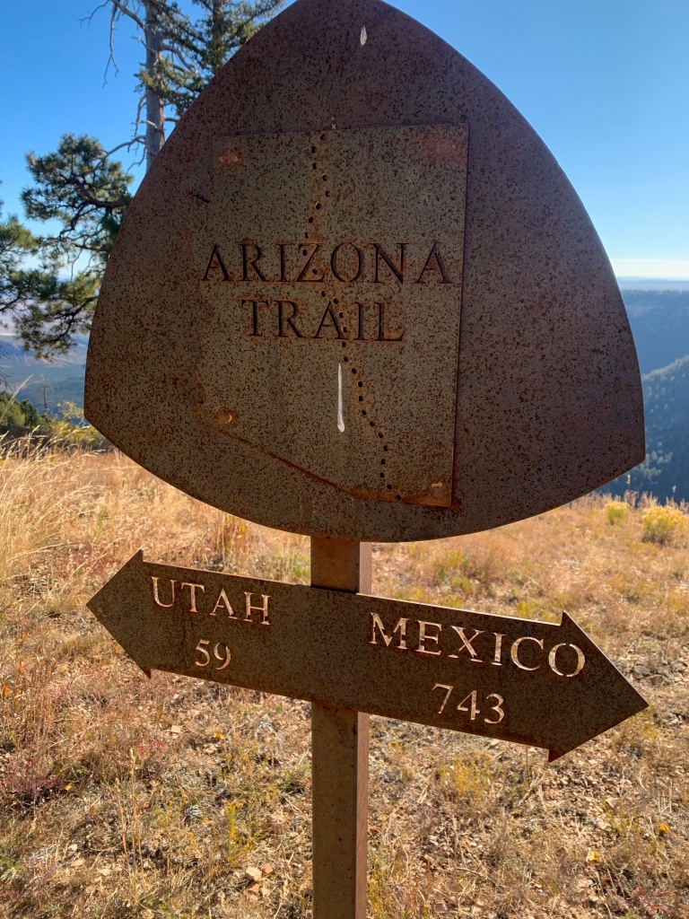 A rusted metal sign holding the Arizona Trail logo stands against the edge of a plateau with arrows pointing in opposite directions.  The arrow to the left reads "Utah 59" and the arrow to the right reads "Mexico 743."