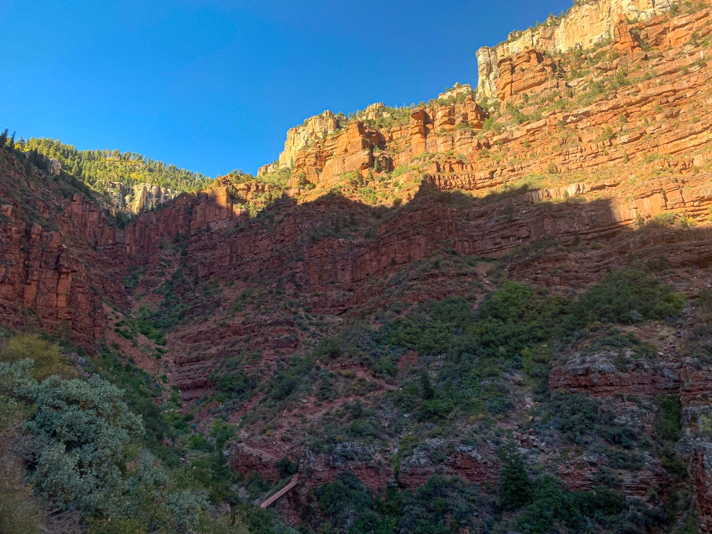 Roaring Springs Canyon split by evening light, with the upper portion glowing red-gold and the lower portion in shadow.  The Redwall Bridge on the Arizona Trail (North Kaibab Trail) across the canyon drainage is visible at the bottom. 