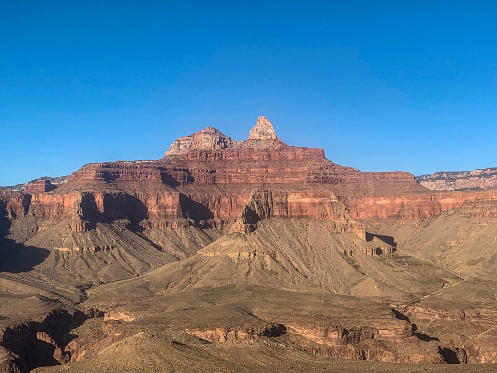 A red ridgeline with occasional white stripes, a white sawtooth rising above and tan slopes flattening out to a plateau beneath stands against a brilliant blue sky, viewed from the South Kaibab section of the Arizona Trail in Grand Canyon.