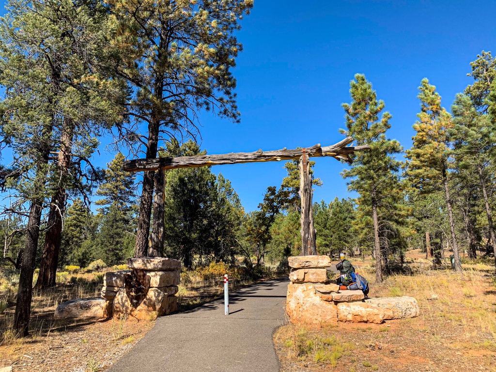 Arizona Trail gateway at the entrance to Grand Canyon National Park.  Two logs standing on top of tan limestone bases support a third log crossing overheat.  A green backpack sits to the side of one stone base as green ponderosa pines stand against a brilliant blue sky in the background.