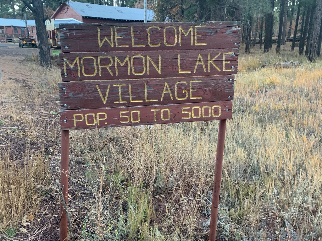 Brown sign with gold lettering reads "Welcome Mormon Lake Village, Pop. 50 to 5000"