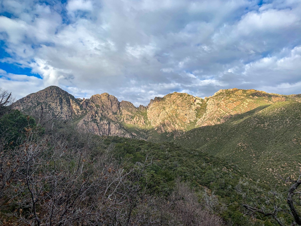 A spot of sun breaks through patchy clouds against a blue sky to shine on a series of four rugged gray mountains with scrubby vegetation on an Arizona Trail thruhike