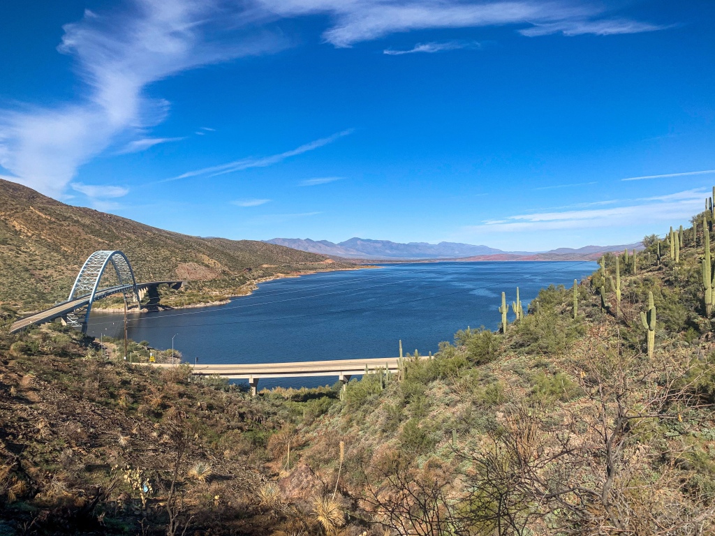 Blue Roosevelt Lake glows beneath greenish brown slopes clad in green saguaro cacti with a blue through-arch bridge to the left and a blue sky above as the Arizona Trail climbs away into the mountains.